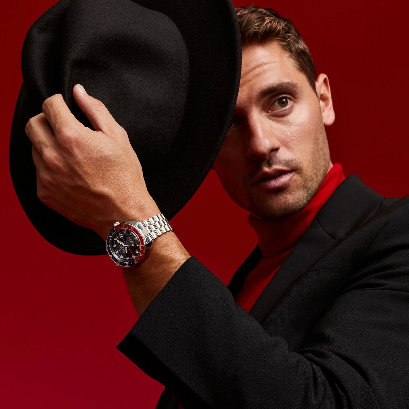 Man met hoed en horloge, rode achtergrond, zwart pak, rode coltrui, poseert