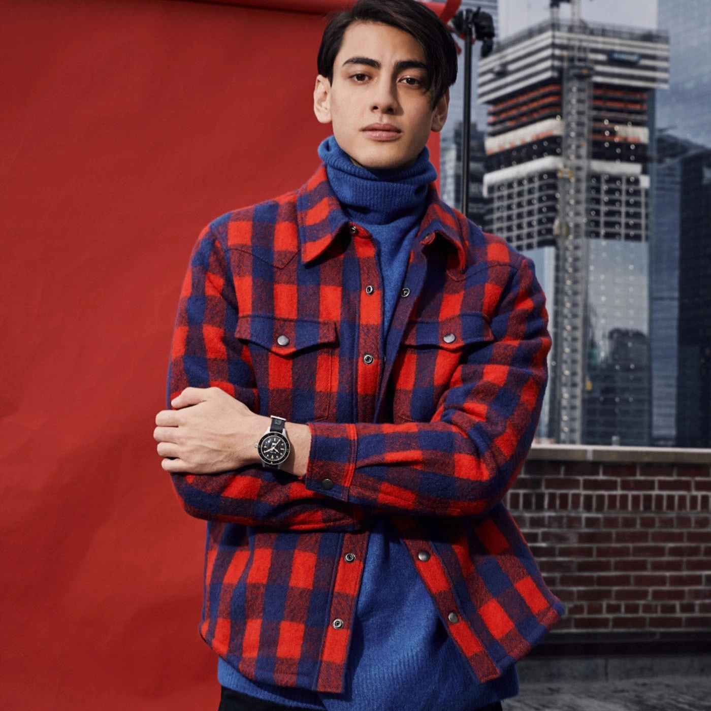 Man in rood en blauw geruit overhemd met een horloge, poseert voor een rode achtergrond