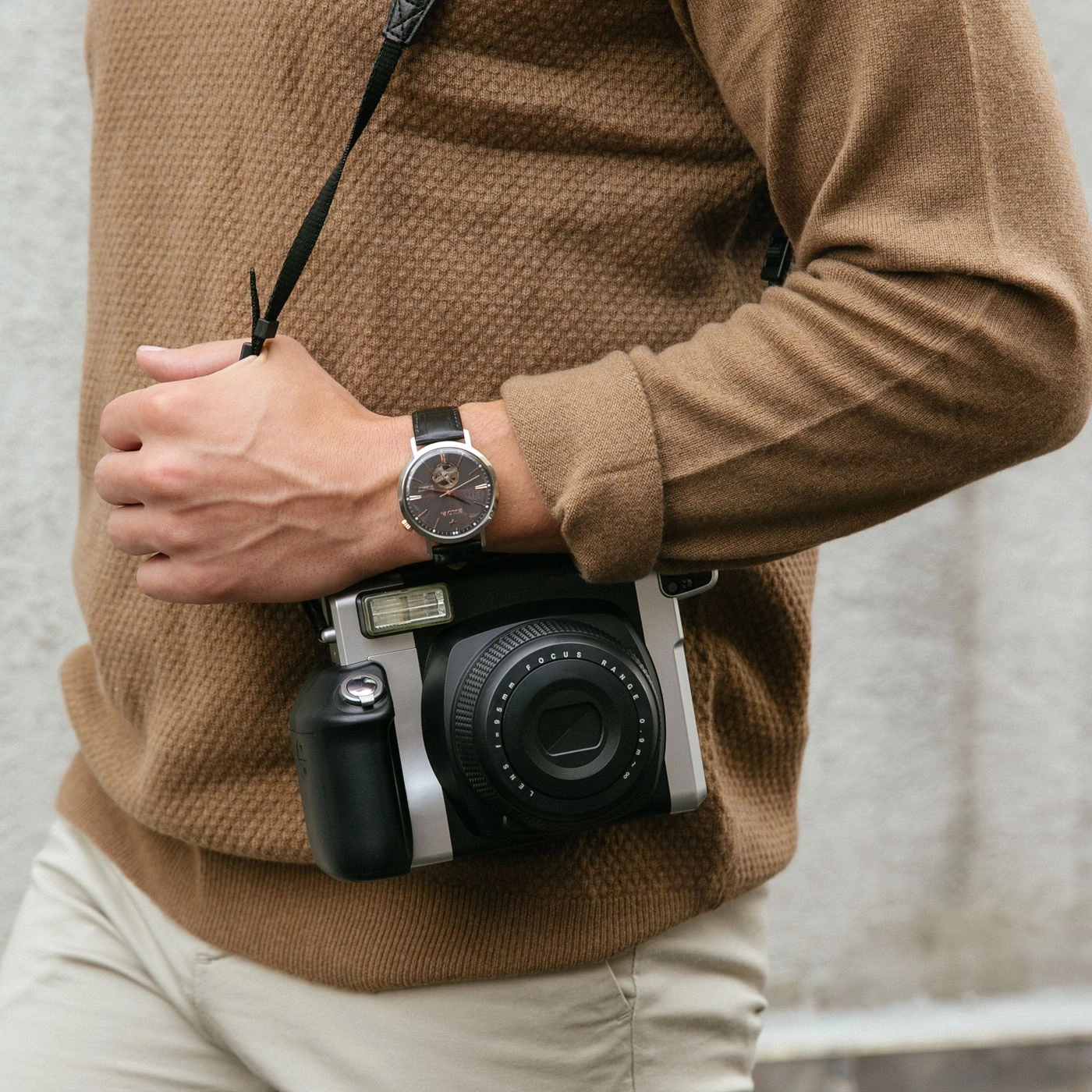 Man met camera en horloge draagt bruine trui en lichte broek.