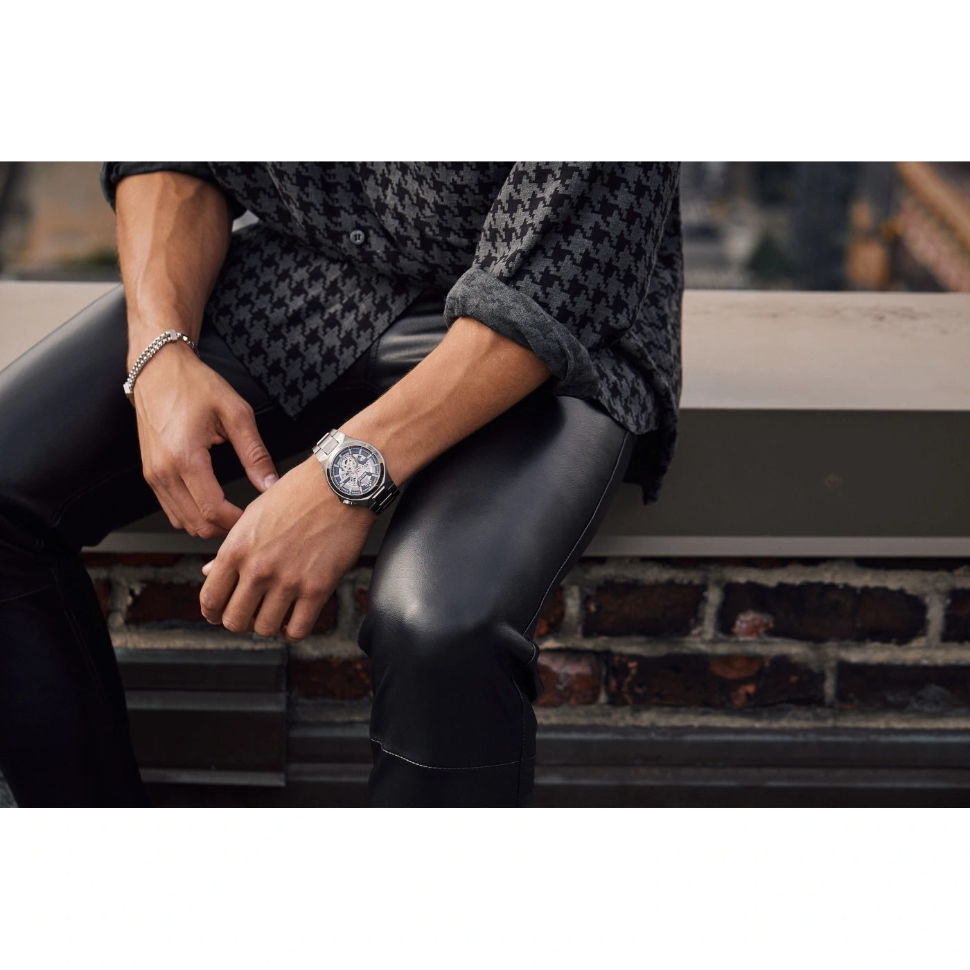 Man met horloge en armband, zittend op muur, leren broek, overhemd