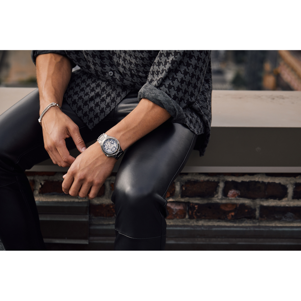 Man met horloge en armband, zittend op muur, leren broek, overhemd